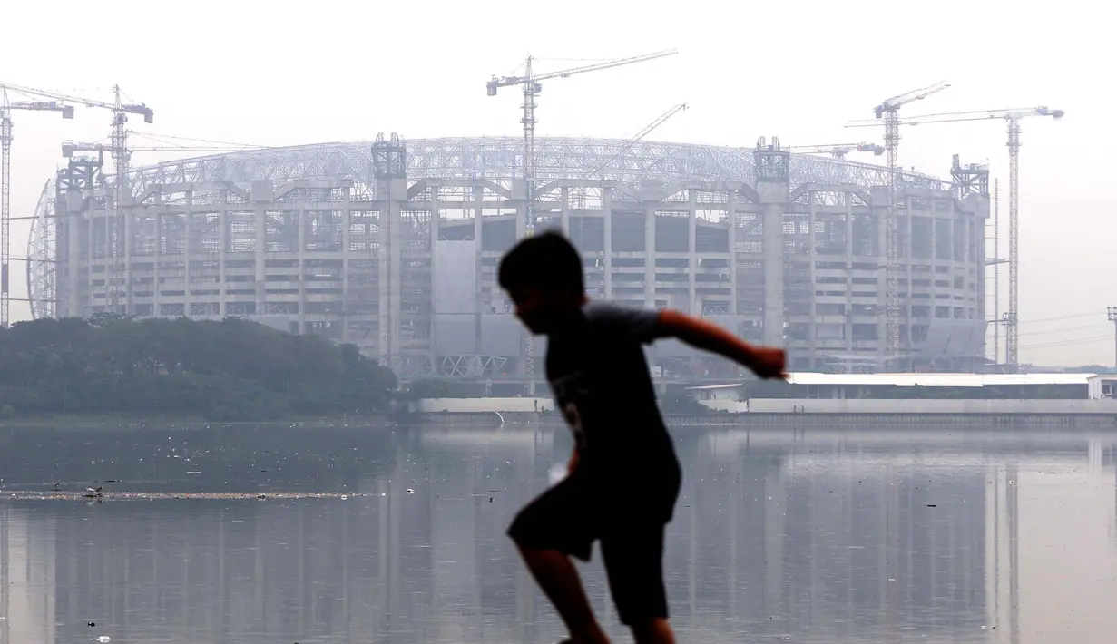 Seorang anak bermain di pinggir Waduk Cincin yang berlatar pembangunan Jakarta International Stadium (JIS), Sabtu (19/6/2021). Pembangunan JIS sudah memasuki tahap pemasangan rangka atap dengan bentang mencapai 269 meter dan ditarget selesai Desember 2021. (Liputan6.com/Helmi Fithriansyah)
