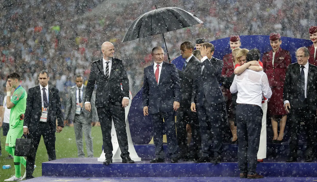 Presiden Rusia, Vladimir Putin dilindungi oleh seorang pemandu yang memegang payung sementara para pejabat lainnya basah kuyup saat hujan turun selama penyerahan medali dan trofi Piala Dunia 2018 di Stadion Luzhniki, Minggu (15/7). (AP/Matthias Schrader)