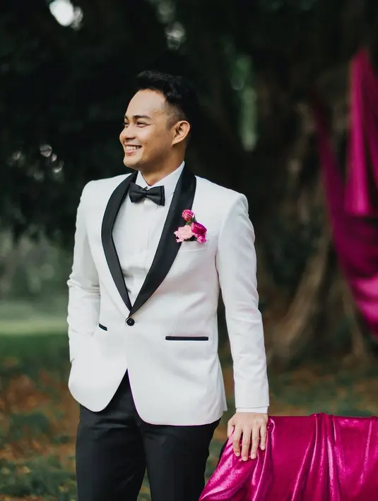 Pasangannya tampil gagah dalam setelan jas putih yang kontras dari @sasdesigns, yang dipermanis dengan bunga-bunga berwarna fuchsia senada, menciptakan harmoni visual antara kedua mempelai. [@terralogical @leurafilm].