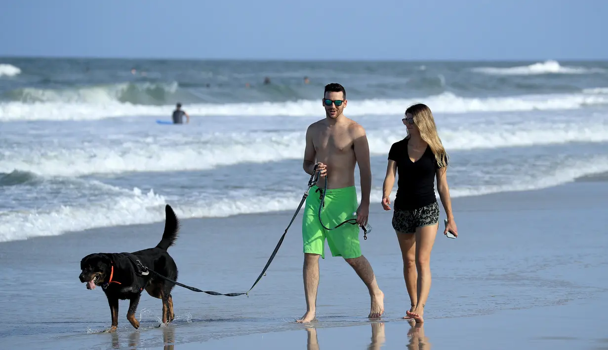 Pasangan membawa seekor anjing berjalan di Jacksonville Beach, Florida (17/4/2020). Pantai dibuka hanya untuk jam terbatas dan hanya bisa digunakan untuk berenang, berlari, berselancar, berjalan, bersepeda, memancing, dan merawat binatang peliharaan. (Sam Greenwood / Getty Images / AFP)