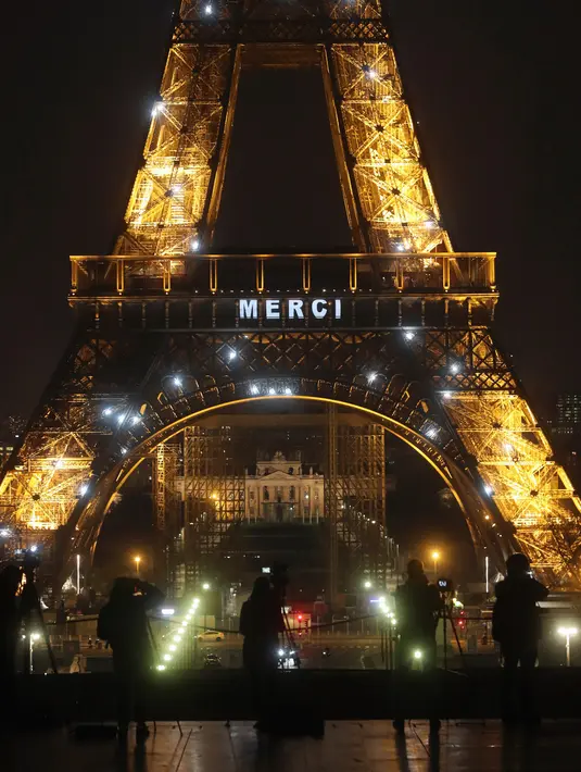Awak media berdiri di depan Menara Eiffel dengan tulisan 'Merci" (terima kasih) yang terpampang di Paris, Jumat (27/3/2020). Menara Eiffel menampilkan pesan dukungan dan terima kasih kepada tenaga kesehatan di Prancis yang berada di garda depan memerangi pandemi COVID-19.  (AP/Thibault Camus)