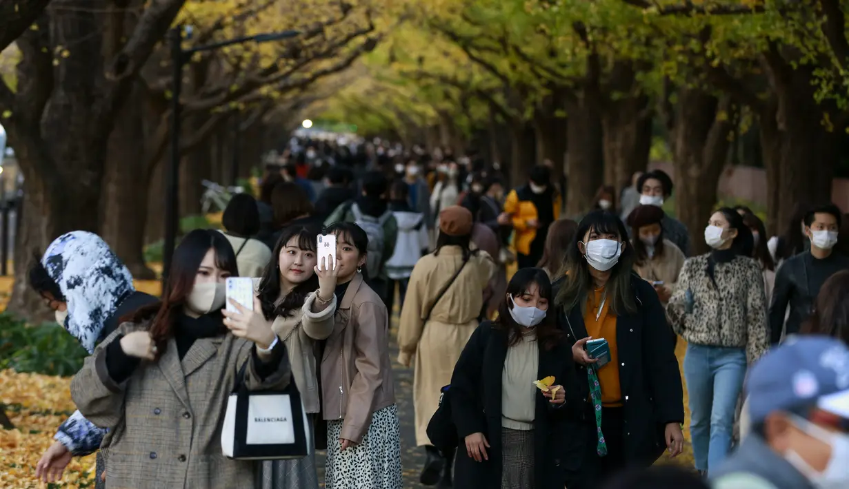 Orang-orang dengan mengenakan masker mengambil foto saat mereka berjalan melalui barisan pohon ginkgo saat pepohonan dan trotoar ditutupi dedaunan kuning cerah di sepanjang trotoar di Tokyo, Jepang pada 28 November 2020. (AP Photo/Kiichiro Sato)