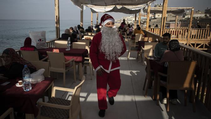 Seorang pramusaji Palestina berpakaian Sinterklas menggendong seorang anak untuk berfoto di sebuah restoran di pantai di Kota Gaza, Minggu (13/12/2020). Restoran tersebut didandani dengan tema Natal, pelayanannya pun berpakaian seperti Sinterklas. (AP Photo/Khalil Hamra)