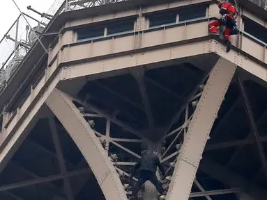 Petugas pemadam kebakaran berusaha mendekati seorang pria (baju hitam di bawah) yang memanjat Menara Eiffel tanpa peralatan keselamatan di Paris, Senin (20/5/2019). Akibat ulahnya, Menara Eiffel ditutup dan pihak pengelola melakukan evakuasi dan pengamanan. (AP/Michel Euler)