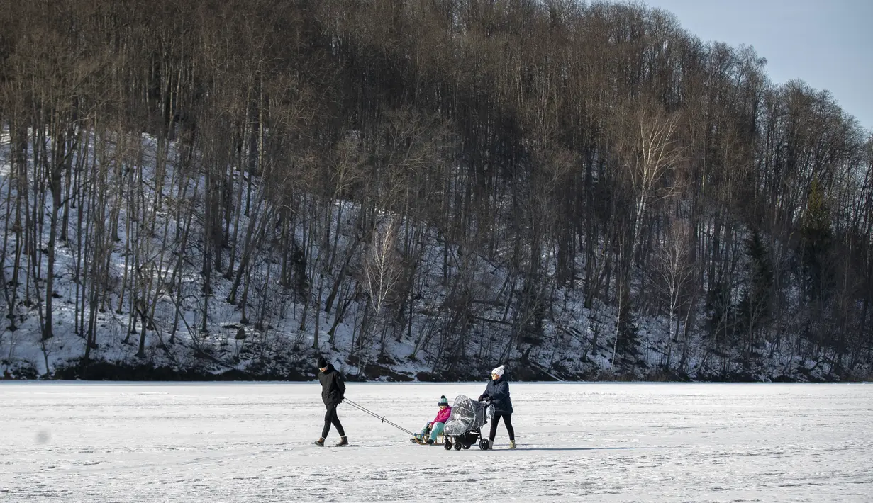 Sebuah keluarga berjalan di danau beku yang tertutup salju dekat Vilnius, Lithuania, Minggu (13/2/2022). Suhu udara 0 derajat Celcius (32 derajat Fahrenheit). (AP Photo/Mindaugas Kulbis)