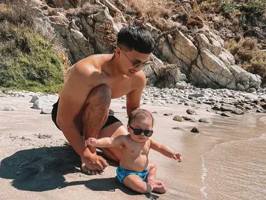 Pelan-pelan, suami Nikita Willy, mengenalkan air kepada anaknya. Sang bayi yang mengenakan kacamata hitam ini didudukan di pasir. (Foto: Instagram/@indpriw)