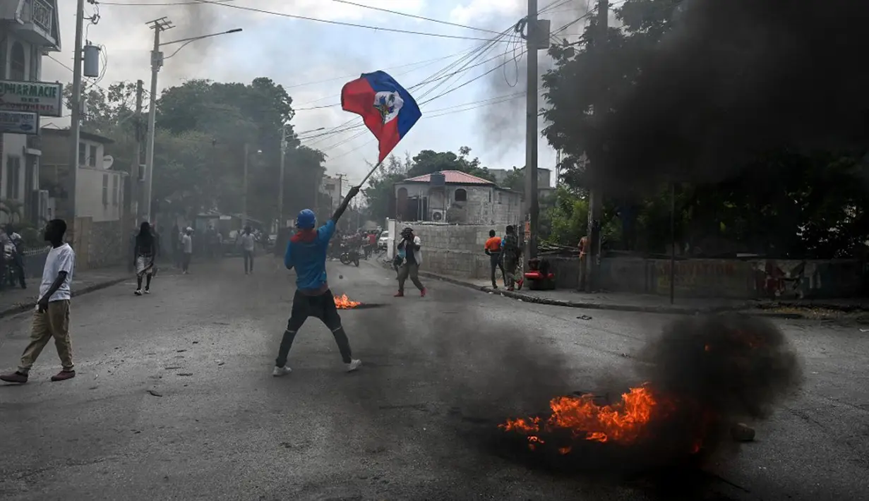 Seorang pria memegang bendera Haiti sementara ban terbakar saat demonstrasi menentang ketidakamanan di Carrefour-Feuilles, Distrik Port-au-Prince, Haiti, 14 Agustus 2023. (Richard PIERRIN/AFP)