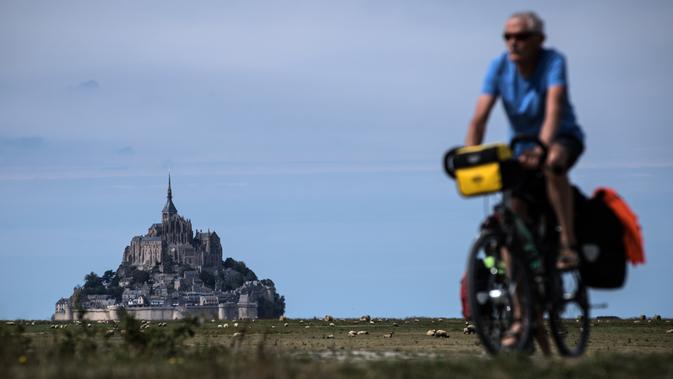 Seorang pria mengendarai sepedanya di sebelah Mont-Saint-Michel di Normandia, Prancis barat laut (31/8/2019). Mont saint Michel atau Pulau Pasang ini selalu dikunjungi sekitar 3 juta wisatawan tiap tahunnya. (AFP Photo/Joel Saget)