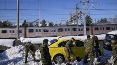Kereta api melintas ketika tentara Yunani mencoba membebaskan taksi yang terjebak di salju, di jalan raya Attiki Odos, setelah hujan salju lebat pada Selasa, di Athena, Rabu (26/1/2022). Upaya tersebut dilakukan ketika pihak berwenang berusaha membersihkan jalan yang terblokir. (AP/Thanassis Stavra