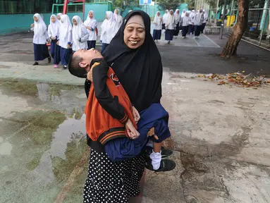 Fauzan Akmal Maulana (15), penderita Distrophia Muculor Progresive (DMP) atau penyakit otot lemah, digendong oleh ibunya, Winih Utami (49), saat bersekolah di SMP Terbuka 1 Tangerang Selatan, pada Jumat (22/3). (merdeka.com/Arie Basuki)
