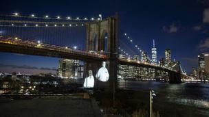 Gambar korban diproyeksikan pada Jembatan Brooklyn saat Hari Peringatan COVID-19 di Brooklyn, New York, Amerika Serikat, 14 Maret 2021. Kota New York yang mencatat kematian tertinggi akibat COVID-19 di AS, memberikan penghormatan yang emosional pada para korban yang meninggal. (Kena Betancur/AFP)