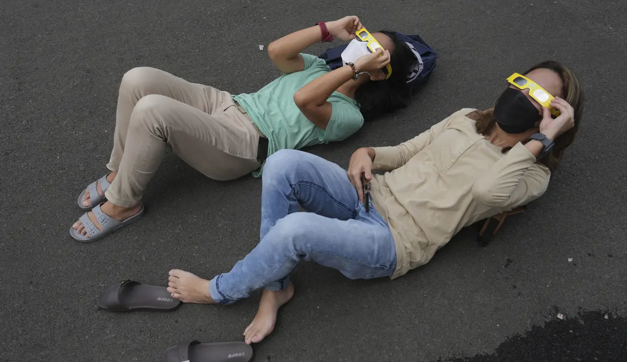 Orang-orang berbaring di tanah saat mereka menggunakan kacamata khusus untuk menyaksikan gerhana matahari hibrida di Jakarta, Kamis (20/4/2023). (AP Photo/Tatan Syuflana)