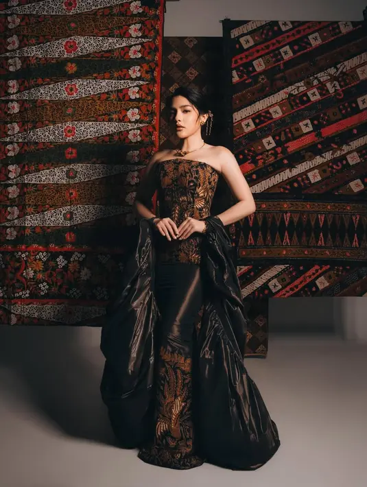 Kain batik yang menjadi aksen utama pada bagian bawah dress memberikan sentuhan tradisional, sementara tambahan outer hitam berukuran besar menciptakan efek dramatis yang begitu kuat. Dipadukan dengan pose editorial dan latar kain tradisional, penampilan Davina terlihat bold, fierce, sekaligus penuh penghormatan terhadap warisan budaya Indonesia. [@davinakaramoy].