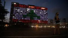 Seorang perempuan Israel yang mengenakan masker berdiri di depan Gedung Balai yang diterangi dengan bendera Lebanon di Tel Aviv, Rabu (5/8/2020). Cahaya bendera Lebanon diproyeksikan sebagai bentuk solidaritas atas ledakan dahsyat di  Beirut yang menghebohkan dunia.  (AP Photo/Sebastian Scheiner)
